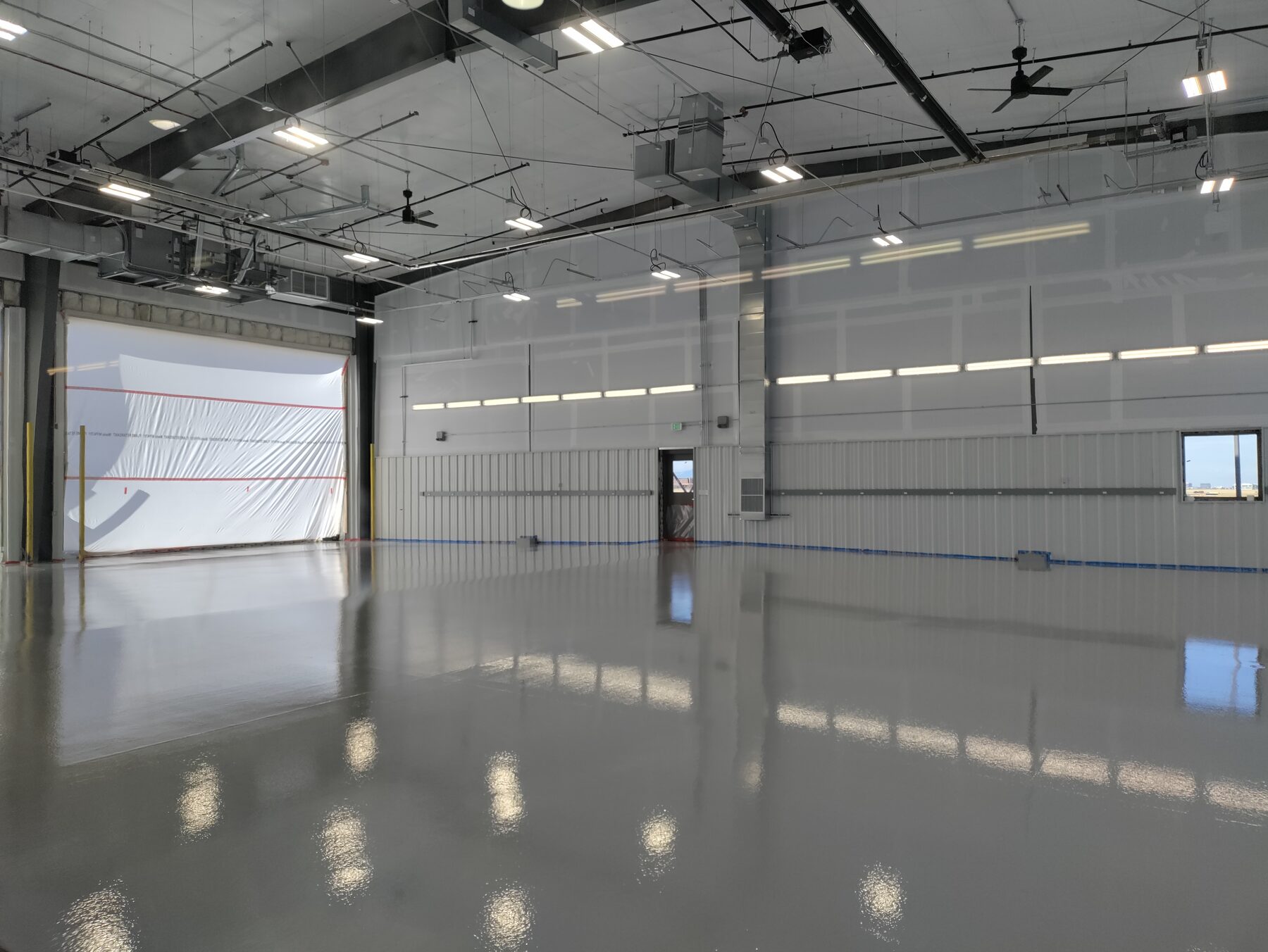 Glossy gray traffic coating across a large empty hangar bay with roll-up door and natural light — traffic coatings Colorado
